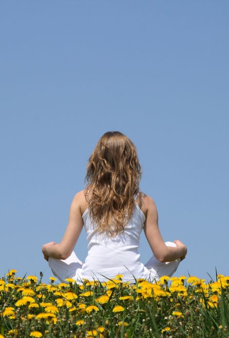 A woman sits in a peaceful lotus pose in a field of wildflowers, her back to the camera, facing a clear blue sky.