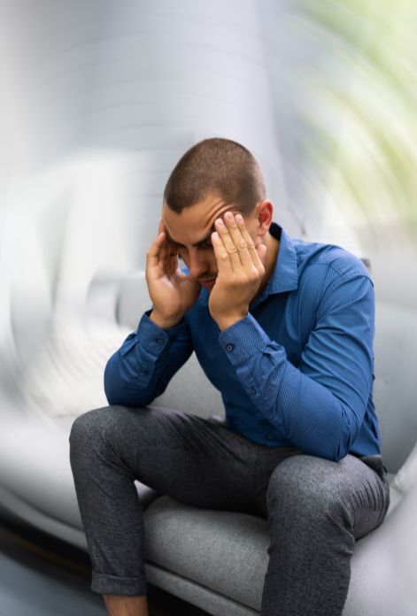 A man sits on a couch, eyes closed, with his hands pressed to his temples in pain. The room around him appears to be spinning, visually depicting the disorienting feeling of brain fog.