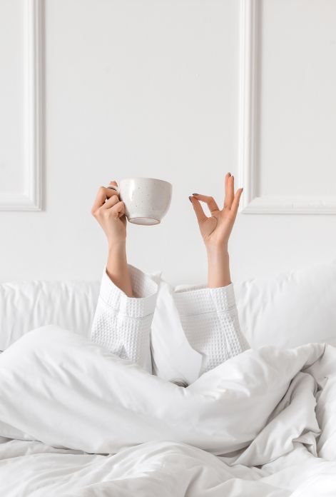 A person lies comfortably in a white bed, surrounded by white linens, holding a white cup and making an "okay" sign, their expression serene and content.