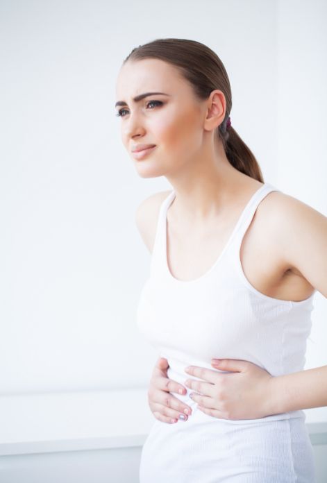 A woman sitting on a bed, hunched over with her hands pressed against her abdomen, her facial expression showing discomfort and pain due to SIBO.