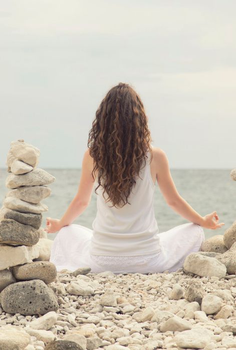 A woman sits in a peaceful lotus position on a beach, facing the ocean with stone formations behind her, symbolizing the tranquility and healing achieved through holistic SIBO treatment at Five Journeys.