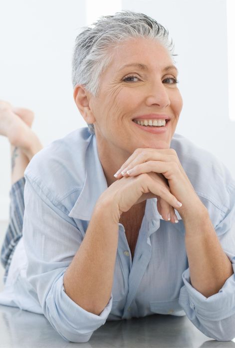 A woman lies on her stomach, propped up on her elbows, with a bright smile and carefree expression, radiating a youthful energy.