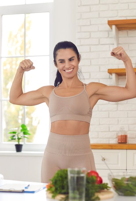 A fit woman flexes her arm muscles with a confident smile in her kitchen, surrounded by an array of healthy foods and green juices, highlighting the connection between nutrition and physical well-being.