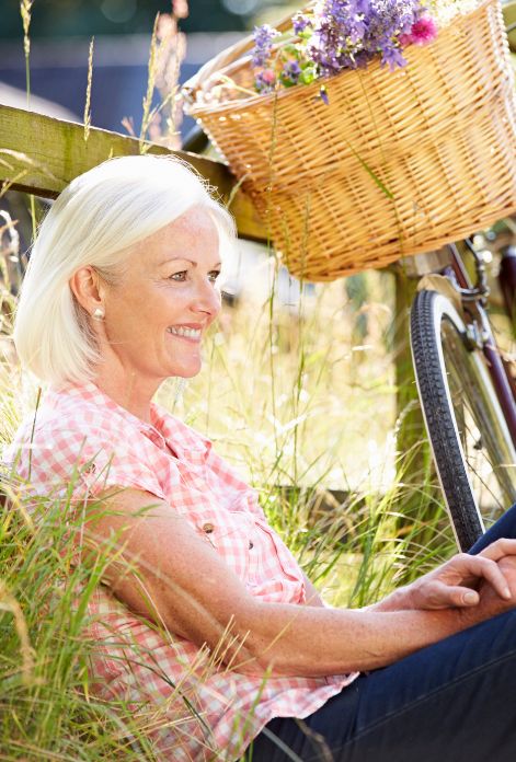 A middle-aged woman with a joyful expression sits on a grassy field, her bicycle with a basket of flowers parked nearby, symbolizing the hope and empowerment that can come with holistic Parkinson's disease treatment.