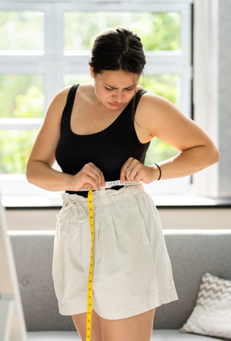 A woman looks frustrated as she measures her waist with a tape measure, reflecting the challenges and emotional toll of resistant weight loss.