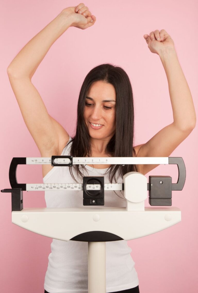 A woman stands on a weighing scale, her arms raised in triumph and a joyful expression on her face, celebrating her success in overcoming weight loss resistance.