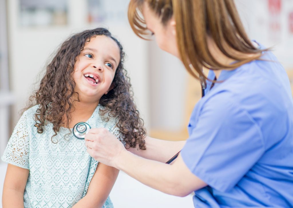 A young girl with a gap-toothed smile beams at a nurse who listens to her heartbeat with a stethoscope, capturing a moment of trust and compassionate care in pediatric medicine.