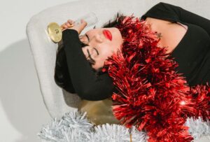 A woman slumps unconscious on a couch, a wine glass loosely held in her hand, amidst festive Christmas decorations, portraying the aftermath of holiday overindulgence.