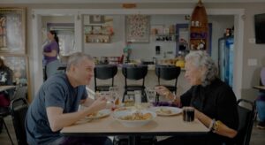 Phil Rosenthal and Chef Sherry share a meal and conversation at a table in the cozy Sly Fox Den Too restaurant, surrounded by eclectic decor and a woman standing in the background.